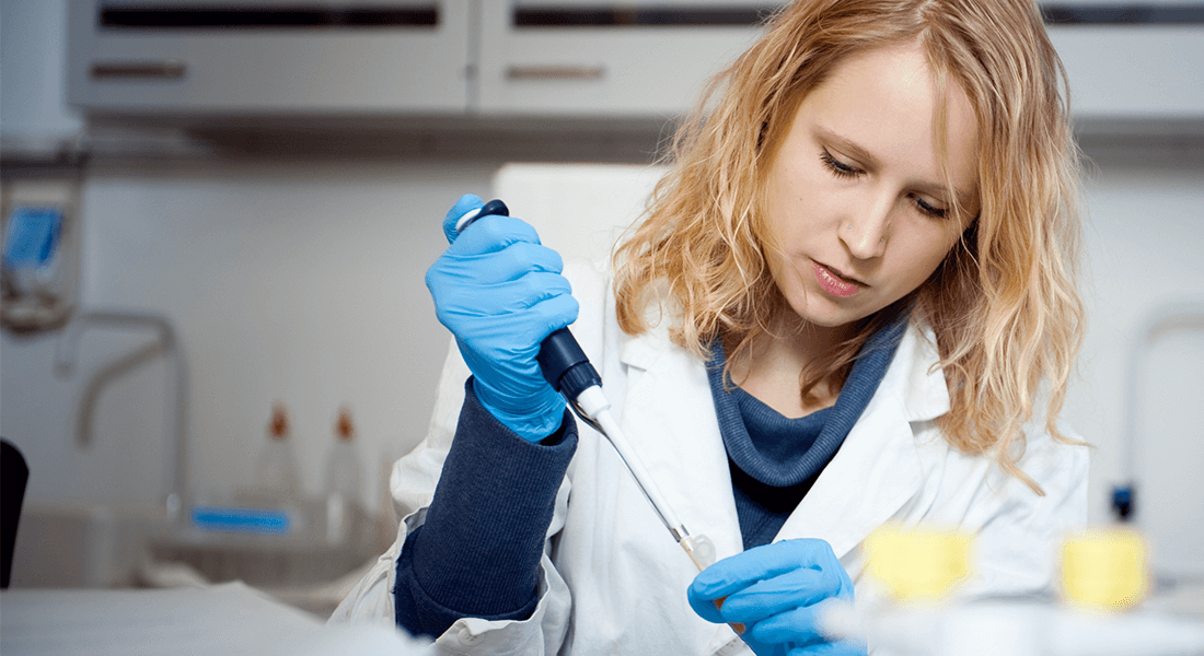 Kvindelig studerende fra Det Sundhedsvidenskabelige Fakultet står med gummihandsker og kittel og er i gang med forsøg